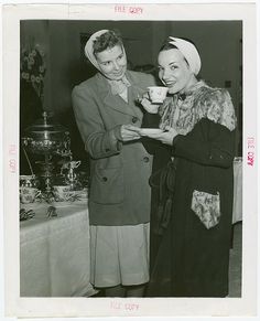 two women standing next to each other in front of a table holding cups and saucers