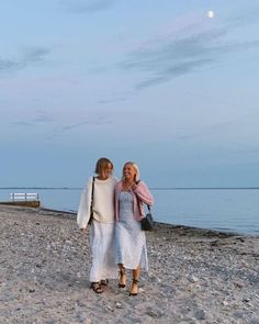 two women are standing on the beach by the water at dusk, one is wearing a white dress and the other has a pink scarf