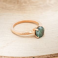 a gold ring with a green stone in it sitting on a wooden surface, next to a piece of wood