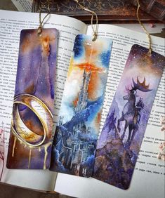 three bookmarks with paintings on them sitting on top of an open book next to a ring