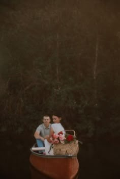 a man and woman sitting in a boat with flowers on the front, surrounded by trees