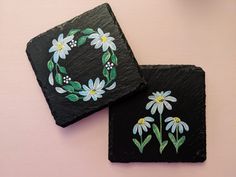 two black slate coasters with painted daisies and leaves on them, sitting on a pink surface