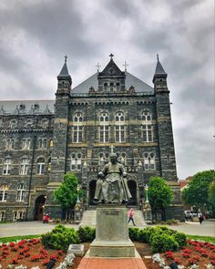 a large building with a statue in front of it and flowers around the perimeter, on a cloudy day