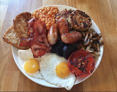 an english breakfast with eggs, bacon, beans and sausage on a white plate sitting on a wooden table