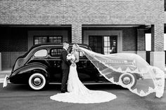 a bride and groom standing in front of an old car with a veil blowing in the wind