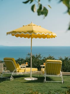 two yellow lawn chairs under an umbrella on the grass by the ocean with tables and chairs in the foreground