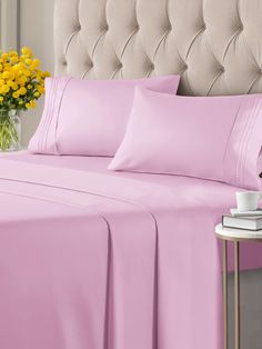 a bed with pink sheets and yellow flowers in the corner next to a white table