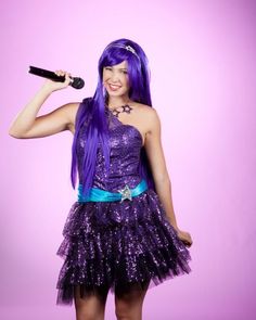 a woman with purple hair is holding a microphone and wearing a purple dress while standing in front of a pink background