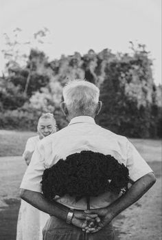 an older man holding a woman in his arms while she looks at the ground with trees behind him