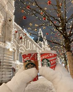 two cups of starbucks coffee in the snow with christmas lights on the trees behind them