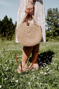 a woman standing in the grass holding a straw bag