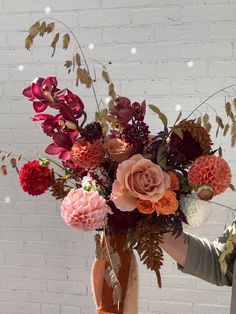 a person holding a vase filled with flowers on top of a wooden table next to a brick wall