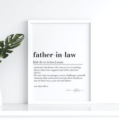 a white vase with a plant in it next to a framed print that says father in law