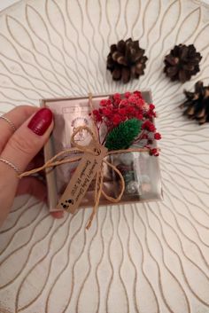 a person is holding a small gift with pine cones and red berries on the table