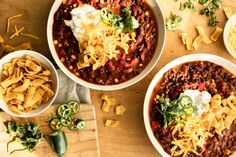 three bowls filled with chili, cheese and tortilla chips on top of a wooden cutting board