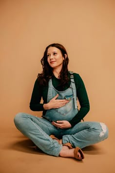 a pregnant woman sitting on the floor with her belly in her hands and wearing overalls