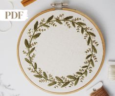 an embroidered wreath with green leaves and sprigs sits on a white surface next to some thread