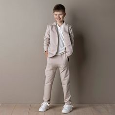 a young boy standing in front of a wall wearing a suit and white shoes with his hands on his hips