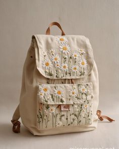 a white backpack with daisies on the front and back pocket, tied to a brown leather strap