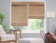 a person holding a remote control in front of a window with roman shades on it