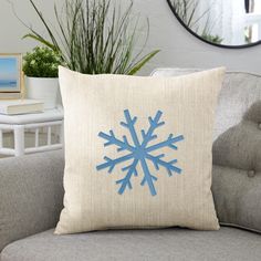 a blue snowflake on a beige pillow sitting on a gray couch next to a potted plant