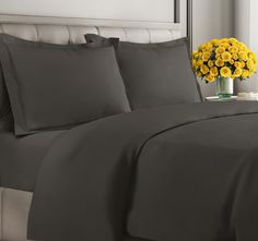 a bed with gray sheets and yellow flowers in a vase on the nightstand next to it