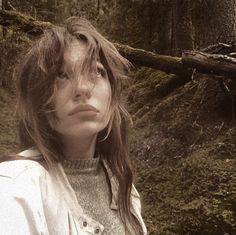 a woman with long hair standing in front of a tree trunk and looking at the camera