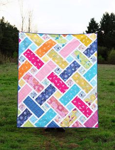 a multicolored quilt sitting on top of a green grass covered field with trees in the background