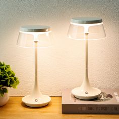 two white lamps sitting on top of a table next to a book and potted plant