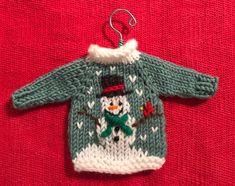 a knitted sweater ornament hanging on a red cloth with a snowman wearing a hat and scarf