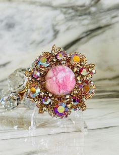 Up for your consideration is this vintage DeLizza & Elster (Juliana) brooch that centerpieces a pink "matrix" cabochon and is surrounded by aurora borealis pink rhinestones and gold tone accent leaves. This D & E brooch also has a pendant bail to adorn a strand of your choice if you prefer to wear it as a necklace.  Unsigned (like all Juliana brooches), but a stunning piece of jewelry history from the 1960s. Condition:  Very good vintage condition, all stones intact and clasp is in wonderful working order. Shows just a bit of wear to the AB coating, but fabulous shape for age. Dimensions:  Approximately 2.25" in diameter FREE SHIPPING via USPS PRIORITY or USPS GROUND ADVANTAGE within the 48 continental states only. All sales are final/no returns... but please contact us if there is an issu Elegant Pink Brooches For Collectors, Vintage Pink Collectible Brooches, Pink Rhinestone Brooches As Gift, Pink Rhinestone Brooches For Gifts, Pink Brooch Jewelry For Wedding, Pink Wedding Brooch Jewelry, Pink Rhinestone Party Brooches, Pink Rhinestone Party Brooch, Pink Party Brooches With Rhinestones