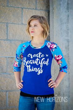 a woman standing in front of a brick wall wearing a blue shirt that says create beautiful things