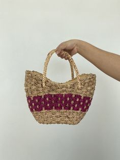 a hand holding a woven straw bag with red and white stripes on the bottom, in front of a white background