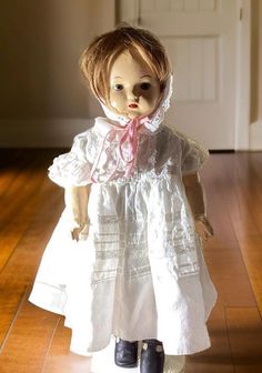 This **unmarked composition doll from the 1930s** stands 20 inches tall and features a composition head, arms, and legs, with a cloth body. She has some cracking on her arms, indicative of her age. The doll is dressed in a white sheer dress adorned with horizontal white lines and lace, complemented by four vertical strips of lace running from the waist to the shoulders. The collar and short, puffy sleeves are also decorated with lace.  She wears a white bonnet with a pink ribbon threaded through White Bonnet, White Sheer Dress, Red Tongue, Boy Cut, Boy Cuts, Sleepy Eyes, White Socks, White Teeth, Red Lip