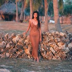 a woman in a long dress standing next to a stone wall with palm trees behind her