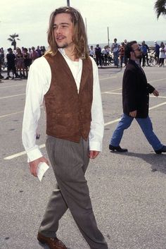 a man with long hair wearing a brown vest and white shirt is walking down the street