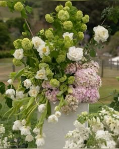 a white vase filled with lots of flowers on top of a lush green field next to trees