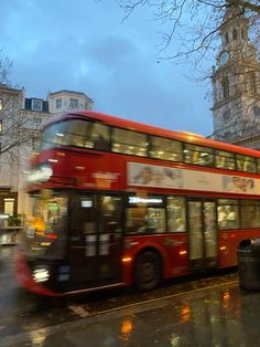 a red double decker bus driving down a street next to tall buildings on a rainy day