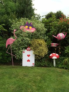 some pink flamingos and other decorations in the grass by bushes with trees behind them