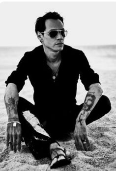 a man sitting in the sand with tattoos on his arms and legs, wearing sunglasses