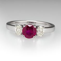 This pretty ring is centered with one (1) round mixed cut natural ruby set into a four-prong setting. The center stone is accented to each side by one (1) oval brilliant cut set into a four-prong setting. The ring measures 5.5mm at the top, rises 5.1mm above the finger, tapering to 2.2mm wide and 1.2mm thick at the base of the shank. This ring is currently a size 6.75. Formal Ruby Three Stone Ring With Diamonds, Luxury Three Stone Ruby Ring, Classic Three Stone Ruby Ring With Diamonds, Luxury Oval Three-stone Ruby Ring, Classic Red Three Stone Diamond Ring, Fine Jewelry Red Diamond Three Stone Ring, Formal White Gold Ruby Ring With Three Stones, Classic Ruby Ring With Center Stone And Round Band, Classic Ruby Ring With Center Stone