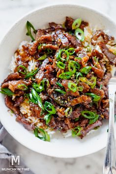 a white bowl filled with meat and rice on top of a marble table next to utensils