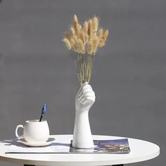 a white vase with some dry grass in it next to a coffee cup and pen