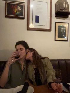 two women sitting at a table with wine glasses in front of their faces, one woman is kissing the other