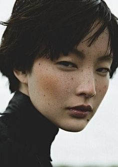 a woman with freckles on her face looking at the camera while standing in front of a body of water