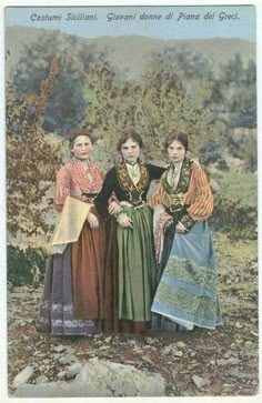 three women standing next to each other in front of some trees and bushes, wearing long dresses