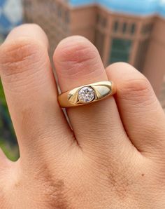 a person's hand holding a gold ring with a diamond in the middle and a building in the background