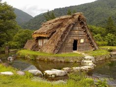 an old thatched house next to a stream in the woods with mountains in the background