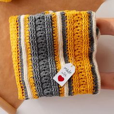 a hand holding a yellow and gray striped crocheted pillow with a red heart on it