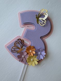 a cake topper with butterflies on it and the number three spelled out in gold, purple, and orange